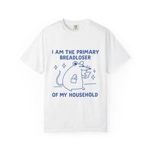 May include: A white short-sleeve t-shirt featuring a blue graphic of a mouse holding a piece of bread. The text reads: I AM THE PRIMARY BREADLOSER OF MY HOUSEHOLD. The design includes small star accents surrounding the mouse illustration.
