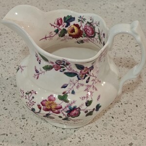 May include: A white ceramic jug featuring a scalloped rim and a curved handle. The exterior and interior rim are decorated with a colourful floral pattern consisting of pink, red, yellow, and purple flowers with green leaves and thin brown stems.