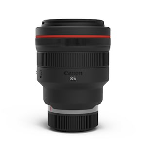 May include: A black Canon 85 camera lens featuring a prominent red ring around the top barrel, textured rubber focus and control rings, and the Canon logo with the number 85 printed in white on the side of the matte black cylindrical body.
