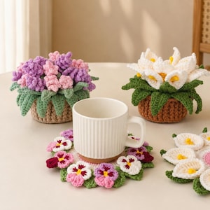 May include: A white ceramic mug with vertical ridges sits on a round crochet coaster featuring purple, pink, and white pansy flowers. Behind it are two crocheted potted plants, one with purple and pink blossoms and one with white calla lilies.