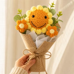 Puede incluir: Un ramo de ganchillo con un gran girasol amarillo con cara sonriente, dos flores naranjas más pequeñas con centros blancos y hojas de eucalipto verdes. Las flores están envueltas en papel de periódico marrón y atadas con cuerda.