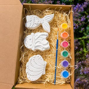 May include: A craft kit in a cardboard box containing three white plaster figures shaped like a mermaid tail, a jellyfish, and a seashell. The set includes a thin paintbrush and a strip of six small paint pots in yellow, orange, pink, green, purple, and blue.