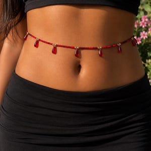 Könnte beinhalten: Eine Taillenkette bestehend aus einem Strang kleiner roter Perlen, die mit goldfarbenen Metallzwischenstücken durchsetzt sind. Die Kette ist mit mehreren hängenden, tropfenförmigen roten Anhängern verziert, die gleichmäßig verteilt sind.
