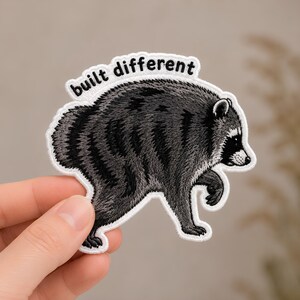 May include: An embroidered iron-on patch featuring a gray and black raccoon in profile, walking forward. The patch has a white border and the text "built different" stitched in black letters in an arc above the raccoon's back.