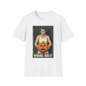 May include: A white short-sleeved t-shirt featuring a graphic print of a person in a white tank top holding a carved orange pumpkin. The illustration includes the text WRONG HOLE! in bold capital letters at the bottom of the rectangular frame.