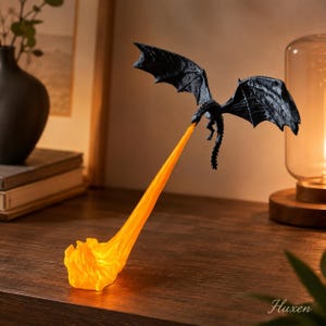 May include: A black dragon figurine in flight, suspended in mid-air, emitting a long, translucent orange flame effect that extends downward to the surface. The dragon features detailed wings and a textured body, creating a dynamic visual display.