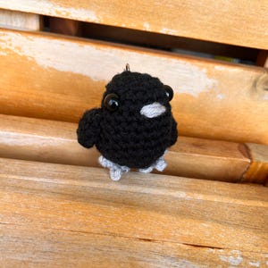 May include: A small, round, black crocheted bird figurine featuring two large, glossy black plastic eyes, a light gray yarn beak, and light gray yarn feet. A small metal loop is attached to the top of the head for hanging as a charm or ornament.