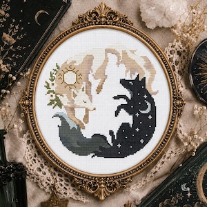 Puede incluir: Un bordado de punto de cruz circular en un marco dorado ornamentado que muestra dos zorros formando un círculo. Un zorro es de color tostado claro con un símbolo solar, el otro es negro con estrellas blancas y una luna creciente sobre tela blanca.