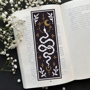 May include: A rectangular cross-stitch bookmark featuring a black background with a white snake coiled in the center. The design includes a gold crescent moon, small gold stars, and white botanical sprigs at the top and bottom corners.