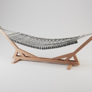Puede incluir: Una hamaca tejida de color gris con flecos decorativos a lo largo de los bordes, suspendida entre dos vigas de soporte de madera clara. La hamaca presenta un diseño de red de malla ancha y está sujeta al marco de madera con correas de tela gris.