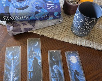 May include: Four rectangular bookmarks featuring blue-toned fantasy illustrations of swords, castles, and figures under moonlight, arranged below two stacked books titled Starside by Alex Aster, a grey ceramic mug, and a small potted succulent.