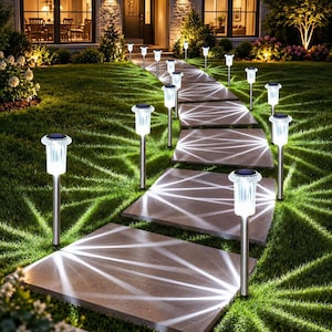 May include: A series of stainless steel solar-powered garden path lights with clear, textured glass shades. Each light is mounted on a thin metal stake and emits a bright white glow, casting distinct, starburst-like patterns across the stone walkway.