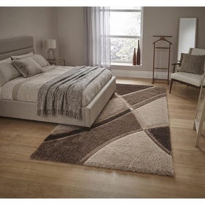 May include: A rectangular area rug featuring a geometric pattern in shades of beige, taupe, and dark brown. The rug has a thick, shaggy pile texture and is positioned on a light-colored wooden floor in a bedroom setting with a bed and furniture.