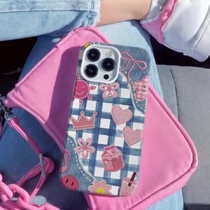 May include: A smartphone case featuring a blue and white checkered pattern overlaid with various pink icons including hearts, a crown, a sun, flowers, a milk carton, and a drink, accented by denim-textured patches at the corners and edges.
