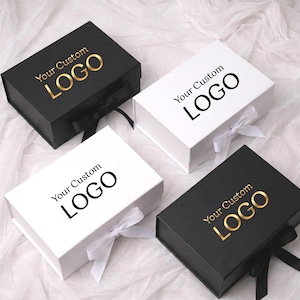 May include: Four rectangular gift boxes with magnetic closures and ribbon ties. Two boxes are matte black with gold foil text reading Your Custom LOGO, and two are white with black text reading Your Custom LOGO. Each box features a matching ribbon bow.