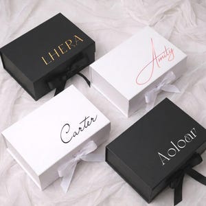 Op de afbeelding: Vier rechthoekige geschenkdozen met magnetische sluitingen en linten. Twee dozen zijn matzwart met gouden en zilveren belettering LHERA en Aoloar. Twee dozen zijn wit met roségouden en zwarte tekst Amity en Carter.