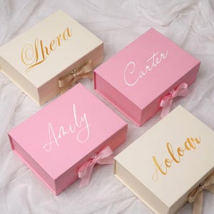 Peut inclure: Quatre boîtes cadeaux rectangulaires avec fermetures magnétiques et rubans. Deux boîtes sont crème avec le texte doré Shera et Aoloar. Deux boîtes sont roses avec le texte blanc Carter et Amily. Les boîtes sont disposées ensemble.