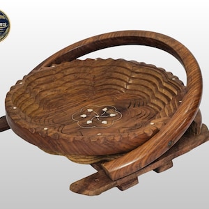 Puede incluir: Una cesta plegable de madera marrón con base circular y asa curva. El interior presenta un patrón floral incrustado con detalles en blanco. La estructura está formada por listones de madera interconectados que permiten plegar la cesta.