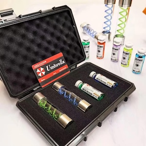 Può includere: Una valigetta protettiva nera con rivestimento in schiuma contenente tre fiale di vetro con tappi in metallo e spirali colorate interne. Una carta rossa e nera con l'etichetta Umbrella Lock System è posizionata all'interno della valigetta.