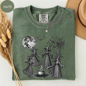 Può includere: Una t-shirt a maniche corte color verde muschio con una stampa grafica che raffigura quattro figure con cappelli a punta riunite attorno a un calderone sotto una luna piena, con pipistrelli e un albero nodoso. Scollo rotondo ed etichetta Comfort Colors.