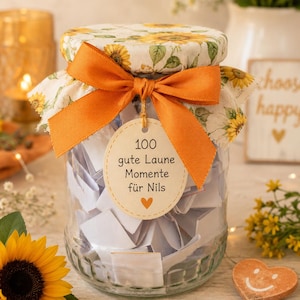 Peut inclure: Un bocal en verre transparent rempli de petits papiers blancs pliés, surmonté d'un couvercle en tissu à motif tournesol et d'un ruban orange. Une étiquette beige ronde est suspendue au ruban avec le texte 100 gute Laune Momente für Nils.