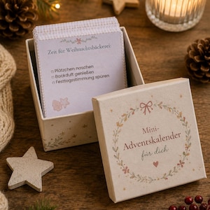 May include: A square cardboard box with a lid featuring a floral wreath design and the text Mini-Adventskalender für dich. Inside, a stack of cards displays the text Zeit für Weihnachtsbäckerei, Plätzchen naschen, Backduft genießen, and Festtagsstimmung spüren.