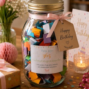 Könnte beinhalten: Ein klares Glas mit goldenem Deckel, gefüllt mit bunten gefalteten Zetteln. Ein hellrosa Band befestigt ein braunes Kraftpapier-Etikett mit der Aufschrift Happy Birthday am Hals. Ein Etikett vorne trägt die Aufschrift Tiny Joys 365 Geburtstagswünsche.