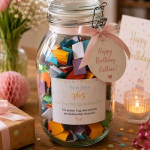 Könnte beinhalten: Ein durchsichtiges Glas mit Bügelverschluss, gefüllt mit kleinen, gefalteten bunten Zetteln. Ein rosa Band ist um den Hals gebunden, mit einem runden Anhänger mit der Aufschrift Happy Birthday, Kathrin! Das Etikett trägt den Text Tiny Joys 365 Geburtstagswünsche.