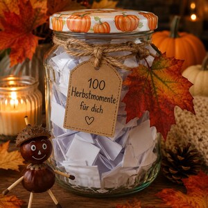 May include: A clear glass jar filled with folded white paper slips, featuring a lid decorated with orange pumpkins. A brown paper tag tied with twine reads 100 Herbstmomente für dich with a heart symbol. A chestnut figure stands beside the jar.
