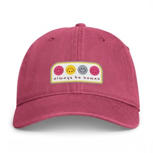 May include: A dark pink baseball cap featuring a rectangular embroidered patch on the front. The patch displays four small smiley faces in pink, yellow, grey, and dark pink, with the text always be human written in brown cursive lettering.