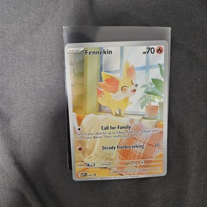 May include: A Fennekin Pokemon trading card featuring a yellow and orange fox-like creature standing on a couch near a window. The card displays 70 HP, the Call for Family and Steady Firebreathing attacks, and text detailing the move mechanics.