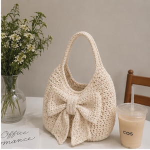 May include: A cream-colored crochet handbag featuring a large, prominent bow attached to the front. The bag has a single integrated handle and a textured, woven stitch pattern throughout its rounded, hobo-style body construction.