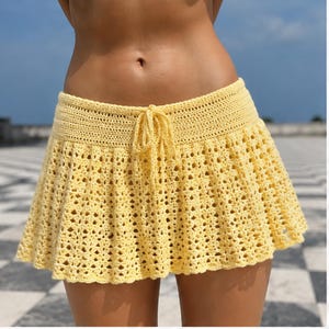 May include: A pale yellow crochet mini skirt featuring a solid waistband with a central drawstring tie and a flared, openwork lace pattern throughout the lower section, ending in a scalloped hemline that creates a textured, lightweight appearance.