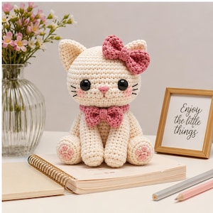 May include: A cream-colored crocheted cat figurine featuring large black eyes, a pink stitched nose, and pink blush on the cheeks. The cat wears a pink crocheted bow on its head and a matching pink bow around its neck, with pink paw pads on its feet.