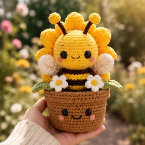 Op de afbeelding: Een gehaakte bijenknuffel met een geel en bruin gestreept lichaam, witte vleugels en een zonnebloemvormig hoofd, zittend in een bruine gehaakte pot. De bij en de pot hebben beide geborduurde zwarte ogen, glimlachen en roze wangen.