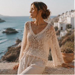 May include: A cream-coloured, long-sleeved crochet jumper featuring an open-knit pattern, a deep V-neckline, and scalloped edges. The garment is paired with light-coloured, loose-fitting trousers that have a drawstring waist detail.