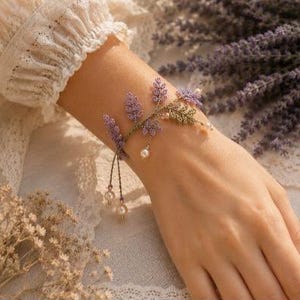 May include: A delicate bracelet featuring intricate purple thread-work shaped like lavender sprigs and small green leaves. The design includes dangling white pearl beads attached to thin green threads, creating a botanical aesthetic on a flexible band.