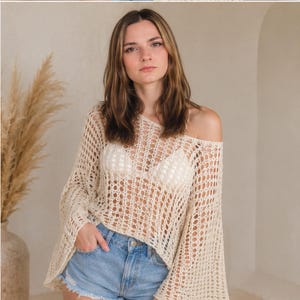 May include: A cream-colored, long-sleeved crochet top featuring an open-knit, grid-like pattern with wide, flared sleeves and a wide neckline, worn over a white camisole, paired with light blue denim shorts with a frayed hem.