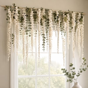 Pode incluir: Um pendente decorativo para janela com múltiplos fios de flores de glicínia em crochê na cor branco sujo, intercalados com folhas de eucalipto de tecido verde, suspensos por uma haste de madeira horizontal sobre a janela.
