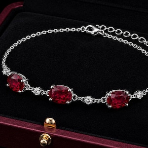 May include: A silver-toned chain bracelet featuring three oval-cut deep red gemstones alternating with small clear round stones. The bracelet includes a lobster claw clasp and an adjustable extender chain, presented inside a dark red jewellery box.