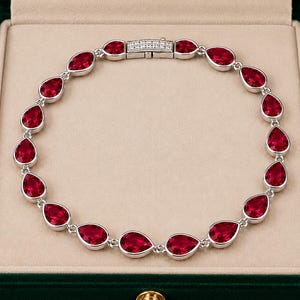 Pode incluir: Uma pulseira de tom prateado com uma série de pedras preciosas vermelho-escuras em forma de pera ligadas entre si. A pulseira inclui um fecho retangular adornado com pequenas pedras claras cravadas em pavé, num design circular.