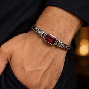 May include: A silver bracelet featuring a woven chain design and a central rectangular deep red gemstone set in a polished silver frame. The metalwork displays intricate detailing at the connection points between the chain and the stone setting.
