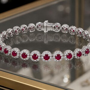 Puede incluir: Una pulsera de tenis en tono plateado que presenta una serie de rubíes redondos de color rojo intenso, cada uno rodeado por un halo de pequeñas piedras blancas transparentes. La pulsera se une mediante un cierre metálico seguro y continuo.