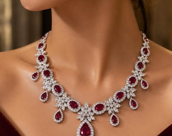 Puede incluir: Un conjunto de collar y pendientes en tono plateado con rubíes de color rojo intenso en forma de pera y corte ovalado, rodeados por halos de pequeñas piedras claras y brillantes. El collar tiene un colgante central.
