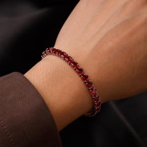 May include: A tennis bracelet featuring a continuous row of oval-cut, deep red gemstones set in a polished silver-toned metal frame. The bracelet encircles the wrist, showcasing a uniform arrangement of faceted stones that reflect light.