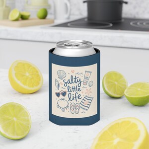 Op de afbeelding: Een donkerblauwe blikkoeler met een crèmekleurig rechthoekig vlak met de tekst "salty little life" in blauw schrift. Het ontwerp bevat illustraties van schelpen, een zonnebril, een tube zonnebrandcrème, een zeester, een handdoek en een armband.