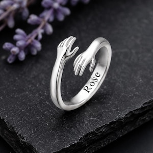 Può includere: Un anello aperto color argento che raffigura due mani che si protendono l'una verso l'altra in un design ad abbraccio. La fascia è incisa con il nome Rose in un carattere serif nero. Il metallo ha una finitura liscia e lucida e una forma curva.