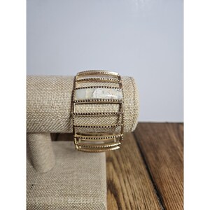 Pode incluir: Uma pulseira tipo bracelete em tom dourado com uma série de barras metálicas horizontais com contas. Uma pedra retangular de cor creme com veios cinzentos está engastada horizontalmente no centro da estrutura de metal vazada, criando um design estruturado.