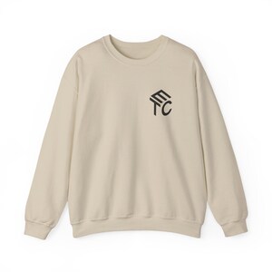 May include: A beige long-sleeve crewneck sweatshirt featuring a black geometric logo on the upper left chest. The logo consists of the letters E, T, and C arranged in a stacked, angular formation. The garment has ribbed cuffs and a ribbed neckline.