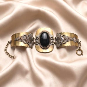 Può includere: Un bracciale con una pietra nera ovale centrale incastonata in una piastra di ottone martellato, affiancata da due pannelli in ottone curvi con dettagli in filigrana color argento. Il pezzo include una catenella di estensione e una chiusura tonda.
